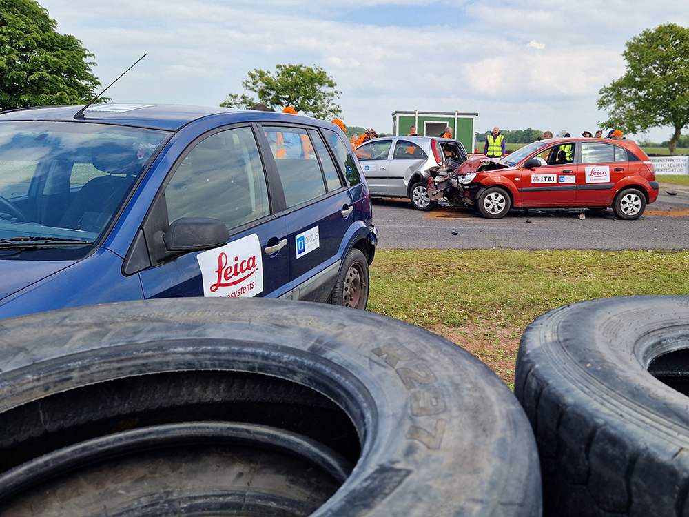 A red Kia with a damaged bonnet and engine bay has crashed into the back of a silver vehicle in the distance. In the foreground, a blue vehicle has collided with the tyre wall of a racetrack.