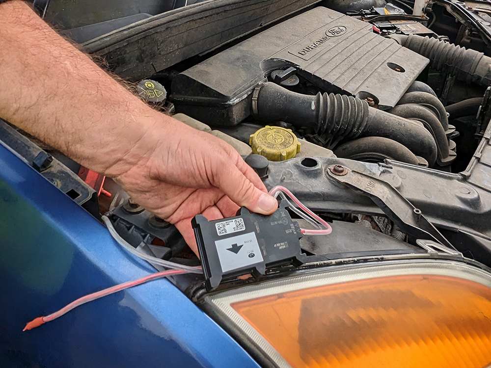 Close up shot of a male hand holding an electrical device over an engine bay of a blue vehicle.
