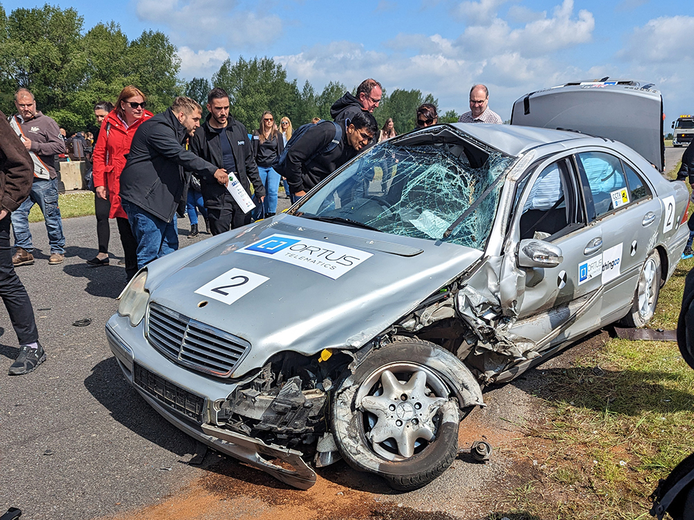 Badly damaged silver car at the side of a racetrack with people gathered round looking at it. The boot is open and the front wheel skewed.