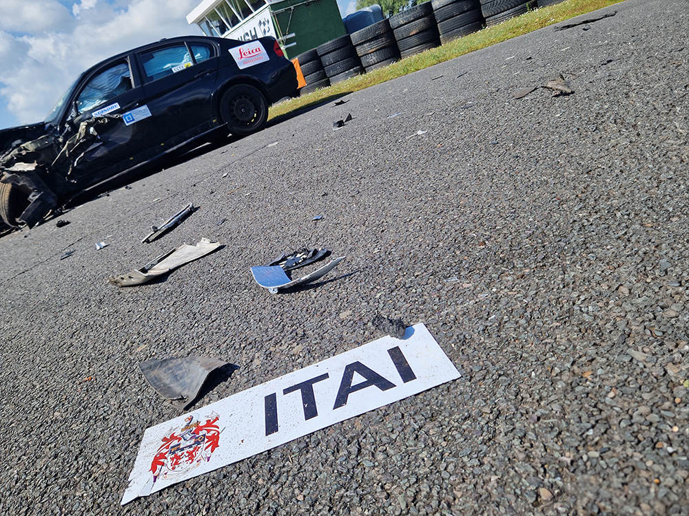ITAI logo on white placque on tarmac, debris leading up to a heavily damaged dark coloured vehicle on a racetrack.