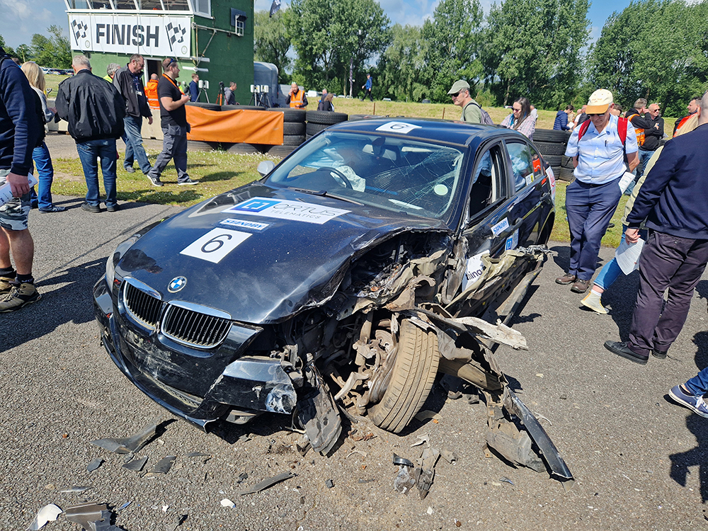 Dark coloured BMW heavily damaged on a racetrack, people crowded round, the passenger side front wheel is skewed and vehicle crumpled. People are gathered around to look.
