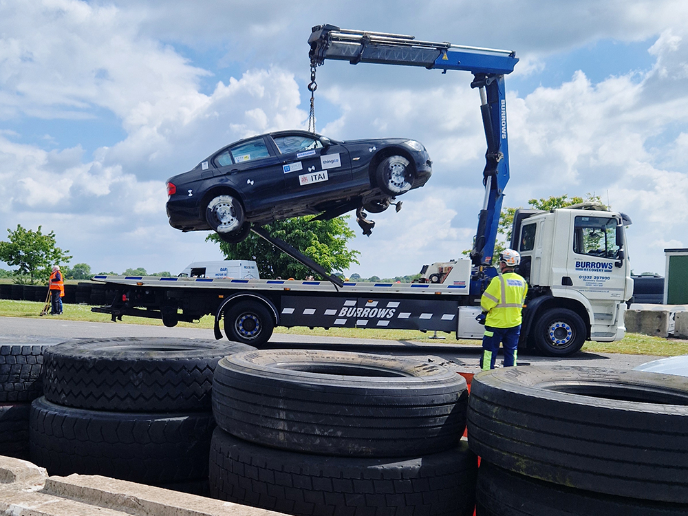 Dark coloured damaged BMW being lifted onto a flatbed Burrows Recovery truck on a racecourse. A tyre wall separates the photographer from the shot.