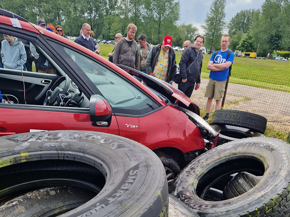 Side view of a heavily damaged red Citroen vehicle which has crashed into a tyre wall at a racetrack. A group of people stand in front looking at it.