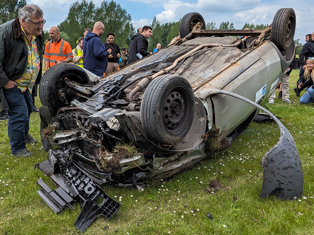 Overturned silver vehicle which is heavily damaged set on the grass, with clumps of grass stuck between the damage. A group of people crowd around to look at it and a man leans in from the lefthand side for a closer look.