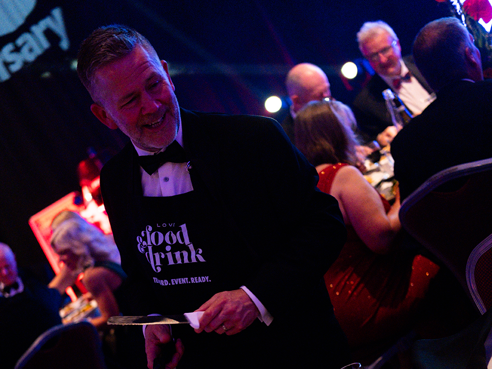 Man in black tuxedo and black apron holds a carving knife smiling at an indoor night time event, other party goers in the background.