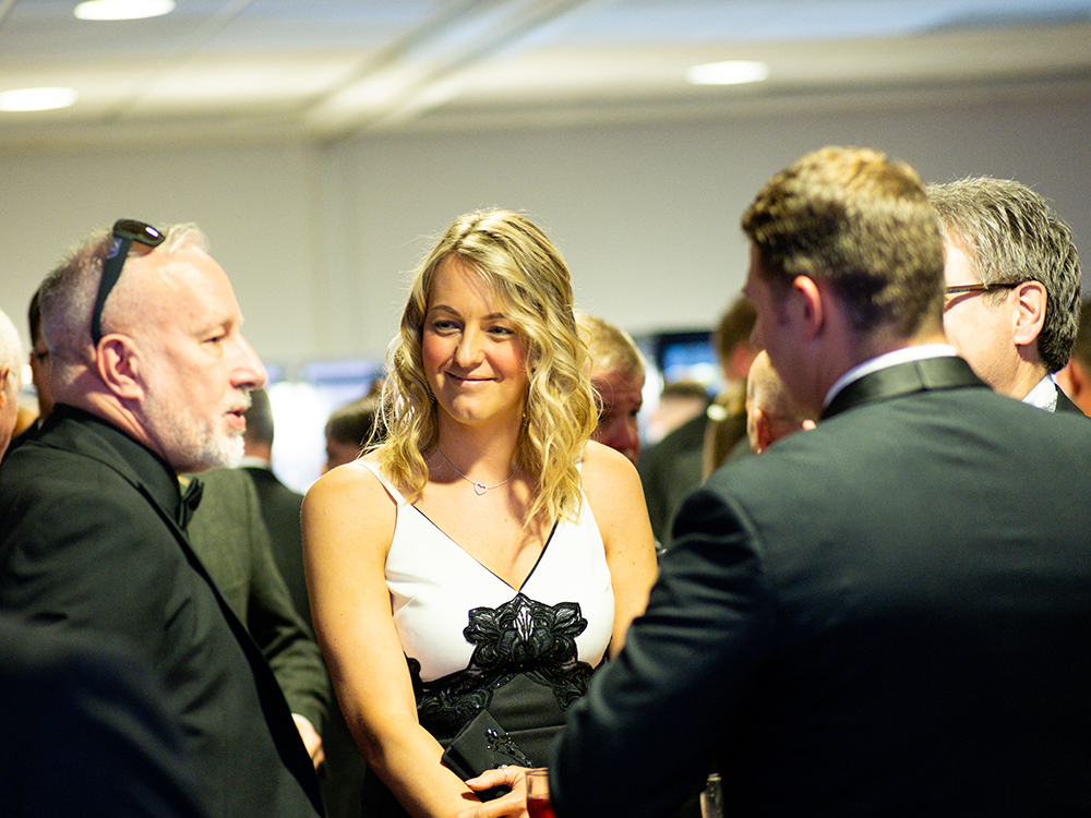 Female in smart evening dress stands between a group of smartly dressed men, smiling and in conversation.