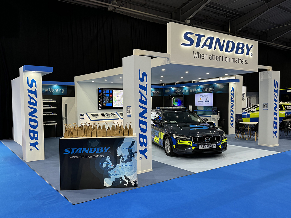 Angle shot from the left of the Standby UK exhibition stand at NAPFM 2024. The stand is white and blue and has an overhanging canopy with LEDs downlighting a Standby branded emergency reponse vehicle. A row of brown paper bags are lined up on the welcome desk.
