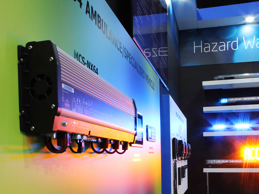 Close up of an MCS-NX64 control unit on an exhibition stand, set on a colourful panel at an angle, leading down to a dark wall with lit up lightbars on white floating shelves. A screen is above the panel with text.
