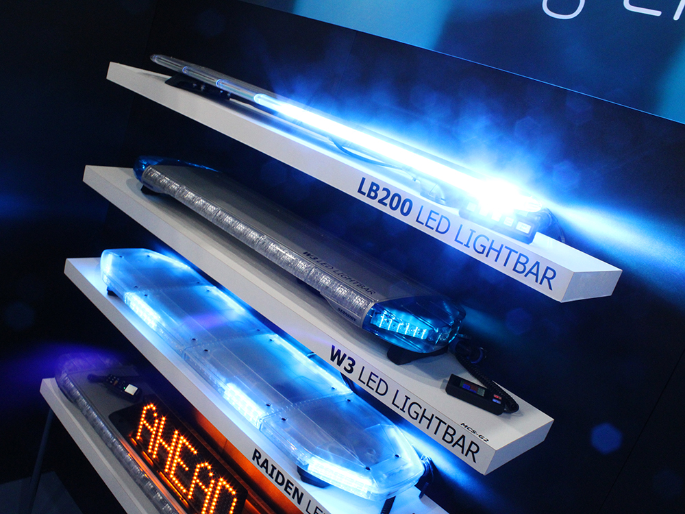 Angle shot of a dark wall on an exhibition stand with white floating shelves and various lit up lightbars on top of each. The topmost lightbar is lit up in blue and has bokeh glare surrounding it. Text on the front of each shelf reads 'LB200 LED Lightbar', 'W3 LED Lightbar' and 'Raiden LED Lightbar'. The bottom lightnar has a scrolling amber matrix display on top, reading 'Ahead'.