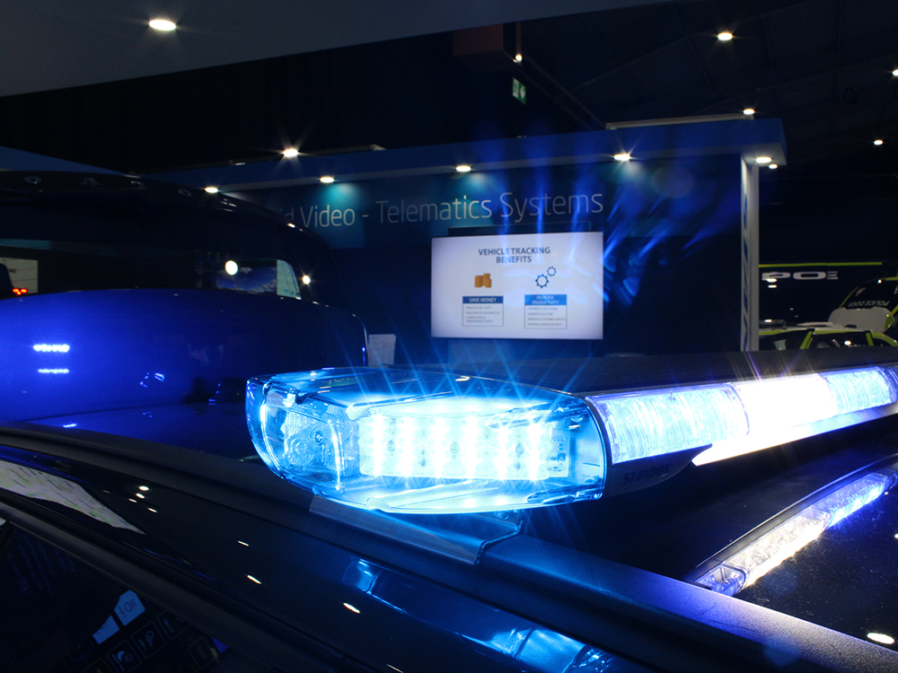 Close up of a blue and white lit lightbar on a dark vehicle roof. The rest of the image is dark, except for a white screen in the background which reads 'Vehicle Tracking Benefits'. The wall behind it reads 'Video - Telematics Systems'.