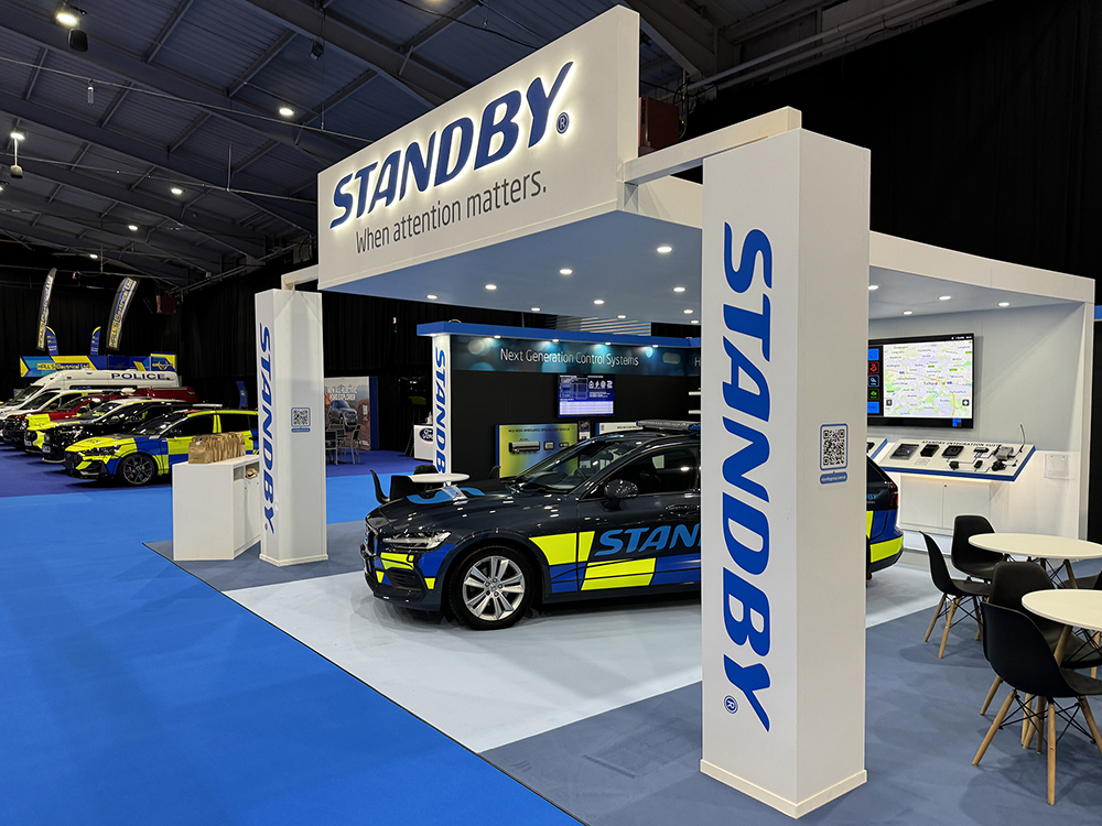 Angle shot from the right of the Standby UK exhibition stand at NAPFM 2024. The stand is white and blue and has an overhanging canopy with LEDs downlighting a Standby branded emergency reponse vehicle. A row of brown paper bags are lined up on the welcome desk.