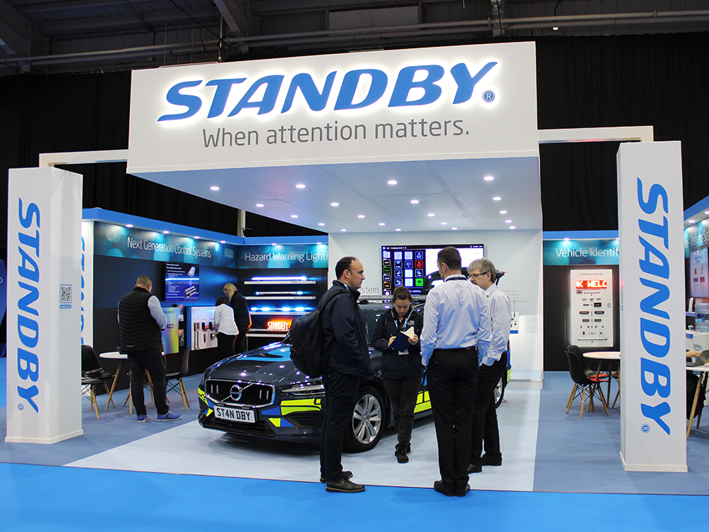 Front shot of the Standby UK exhibition stand at NAPFM 2024. The stand is white and blue and has an overhanging canopy with LEDs downlighting a Standby branded emergency reponse vehicle. Staff and stand visitors are stood around chatting and looking at stand products.