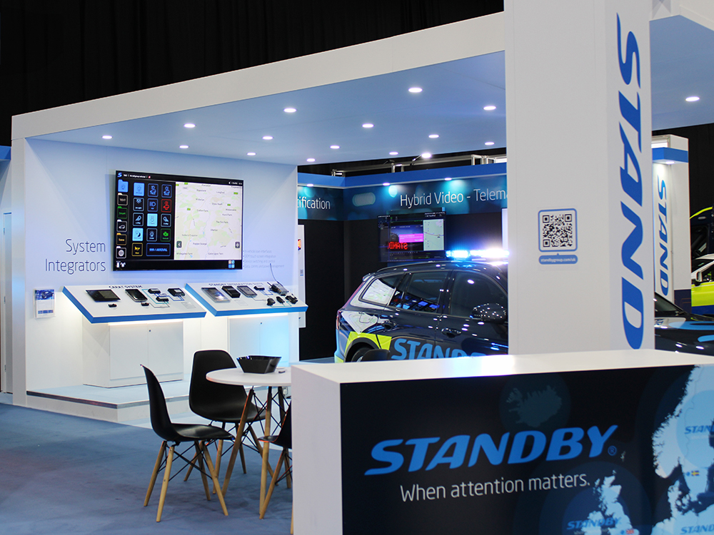 Close up angle shot of the Standby UK exhibition stand at NAPFM 2024. The stand is white and blue and has an overhanging canopy with LEDs downlighting a Standby branded emergency reponse vehicle.