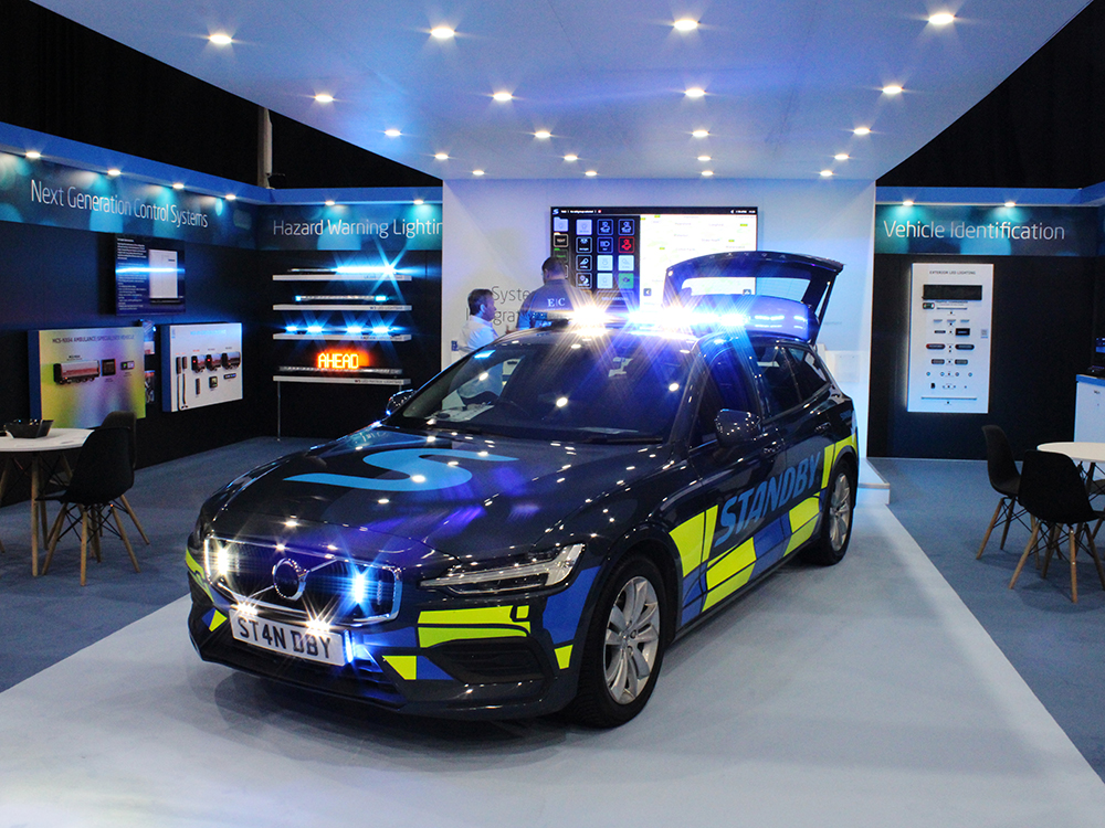Close up shot of a Standby branded emergency response vehicle with blue and white hazard warning lights, parked at an angle under a canopy with LED downlights, on an exhibition stand upon a strip of light blue carpet.