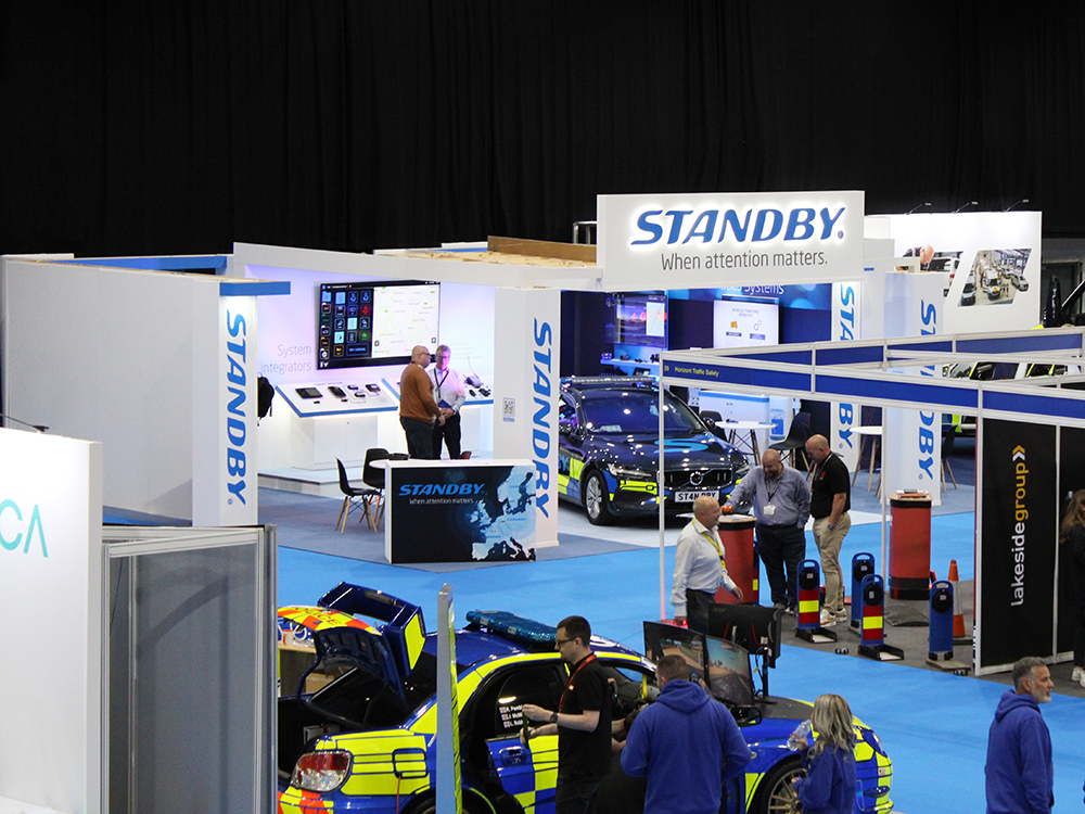Standby exhibition stand in a busy exhibition hall, shot taken from a distance. Compared to other stands in the vicinity, it is more built up. The stand is white and blue and has an emergency response vehicle set under a lit up canopy.