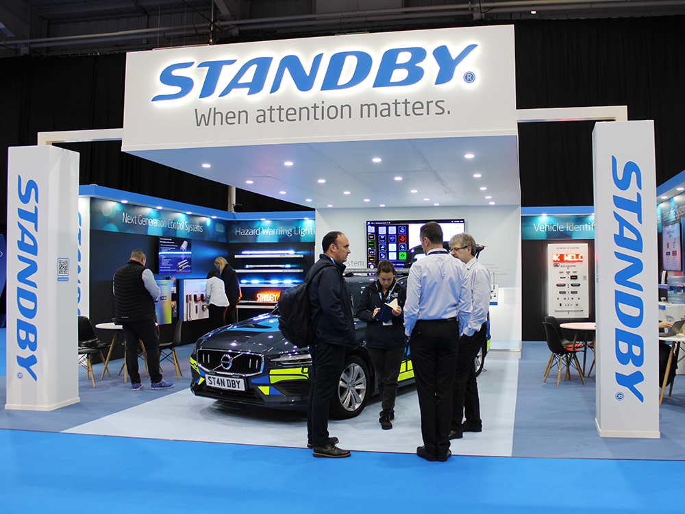 Slightly angled shot of the Standby UK exhibition stand at an indoor event, colleagues and visitors stand around the demo vehicle on the stand, which is lit up by an overhang of LED spotlights, leading to the Standby When Attention Matters signage.