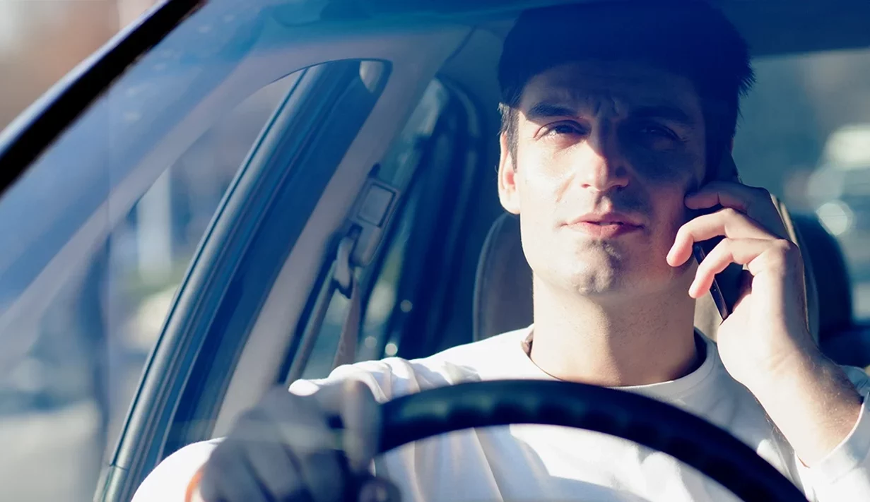 Man in car behind steering wheel on mobile phone taken from in front of the vehicle, closely cropped.