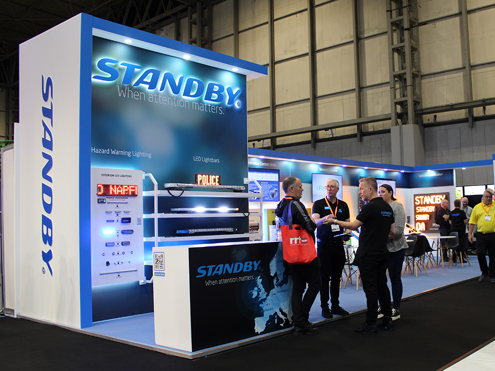 Angle view of Standby UK's exhibition stand at the Emergency Services Show 2024. Two groups of people are on it interacting. The stand is modern and well light, with various pieces of emergency lighting equipment lit in blue and amber.