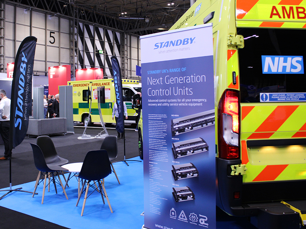 Rear angle view of an ambulance next to a pop-up banner which reads 'Standby UK's range of Next Generation Control Units' with information and images of the product. Four black chairs surround a table on the lefthand side, next to a black flag banner with the Standby logo.