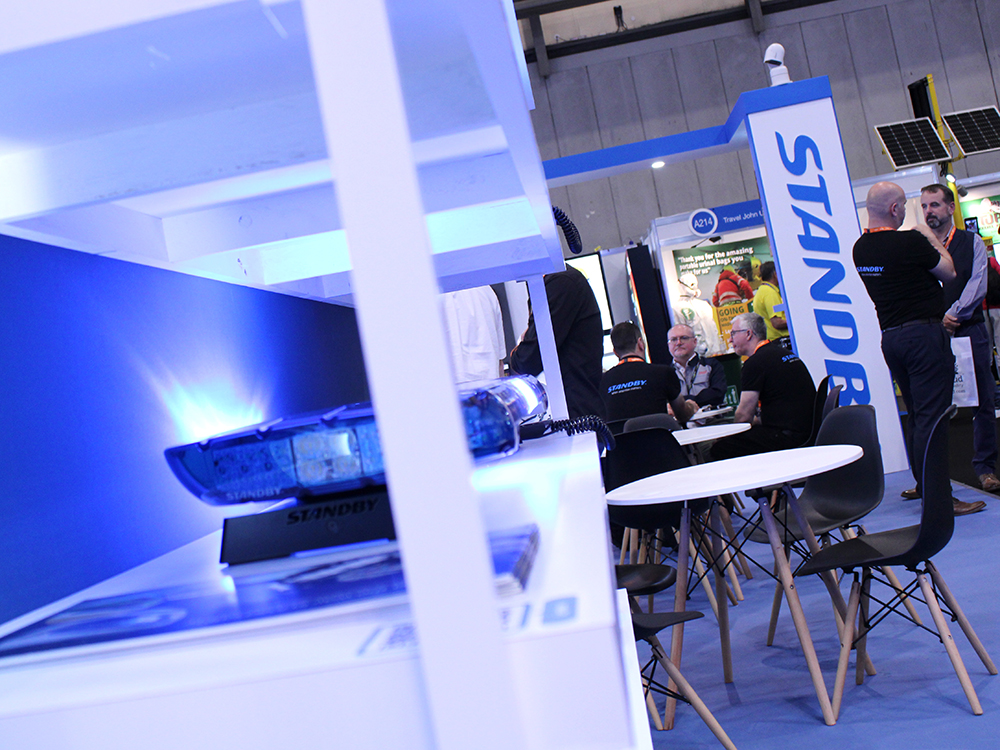 Angle view of the Standby UK exhibition stand at the Emergency Services Show 2024. The image is taken from a viewpoint behind shelves with a lightbar on and two groups of people can be seen interacting, one sitting and one standing.