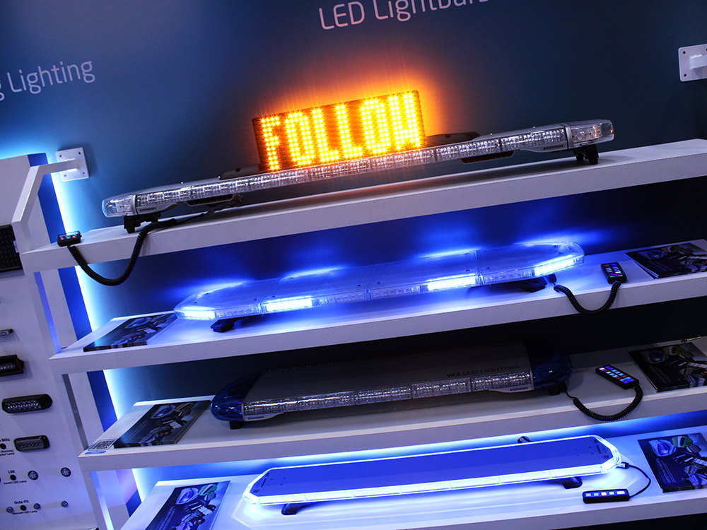 Angle view of a white shelving unit against a dark background with various lightbars on display lit up in blue. The topmost lightbar has an amber LED matrix sign mounted on it which reads 'Follow'.