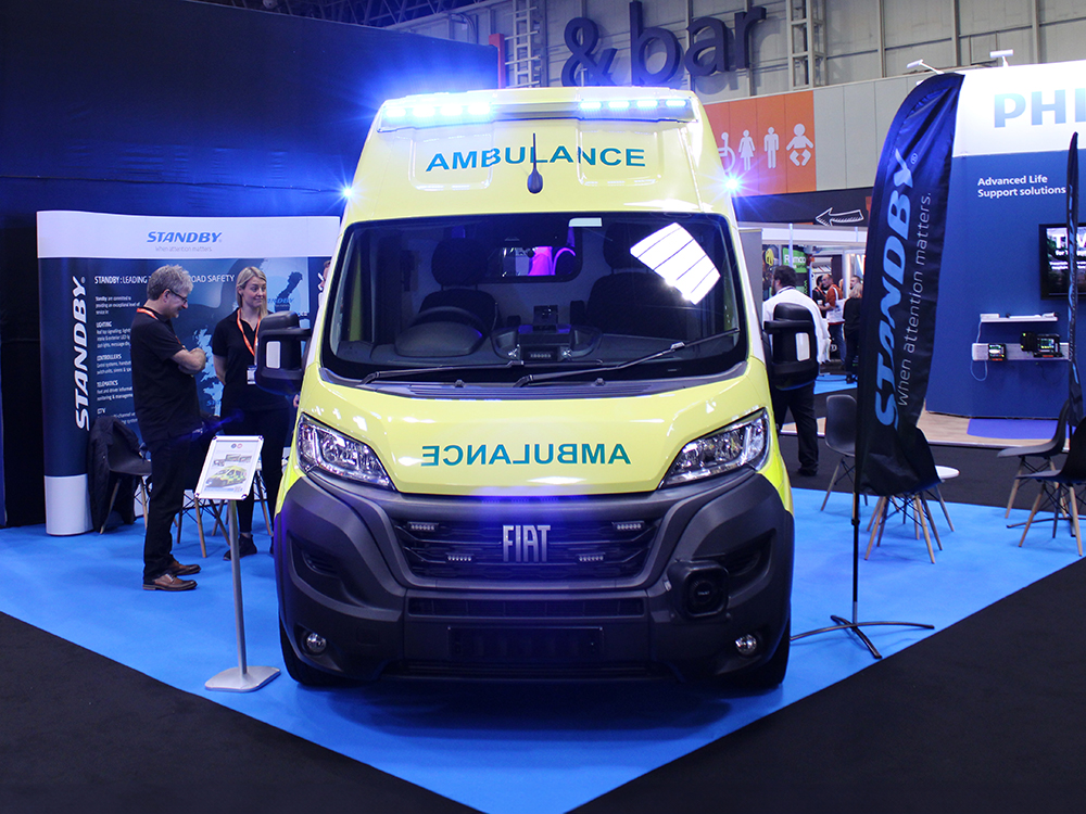 Front view of an ambulance on an exhibition stand at the Emergency Services Show 2024. A group of people stand to it's left in front of a banner which reads 'Standby'. There is also a black flag to the right with the same Standby logo.