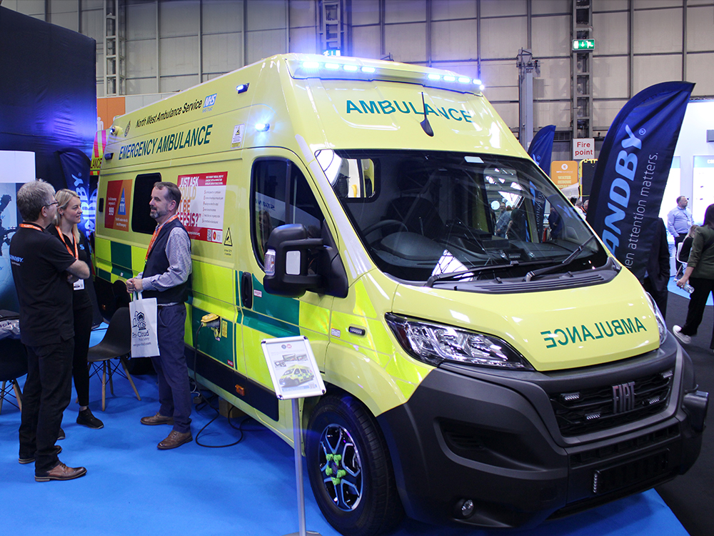 Front angle view of an ambulance on an exhibition stand at the Emergency Services Show 2024. A group of people stand to it's left in front of a banner which reads 'Standby'. There is also a black flag to the right with the same Standby logo.