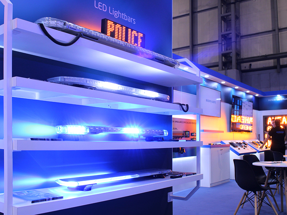 Closely cropped angle view of a white shelving unit with an open back against a dark stand graphic, each with a blue lightbar lit up, shown on a modern looking exhibition stand at the Emergency Services Show 2024.