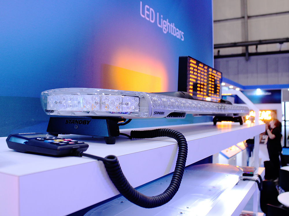 Closely cropped angle view of an unlit lightbar on a white shelf with an amber LED matrix display mounted on top of it. The dark stand graphic behind it reads 'LED Lightbars' and the matrix sign reads 'Police'. The rest of the exhibition stand is out of focus in the background.