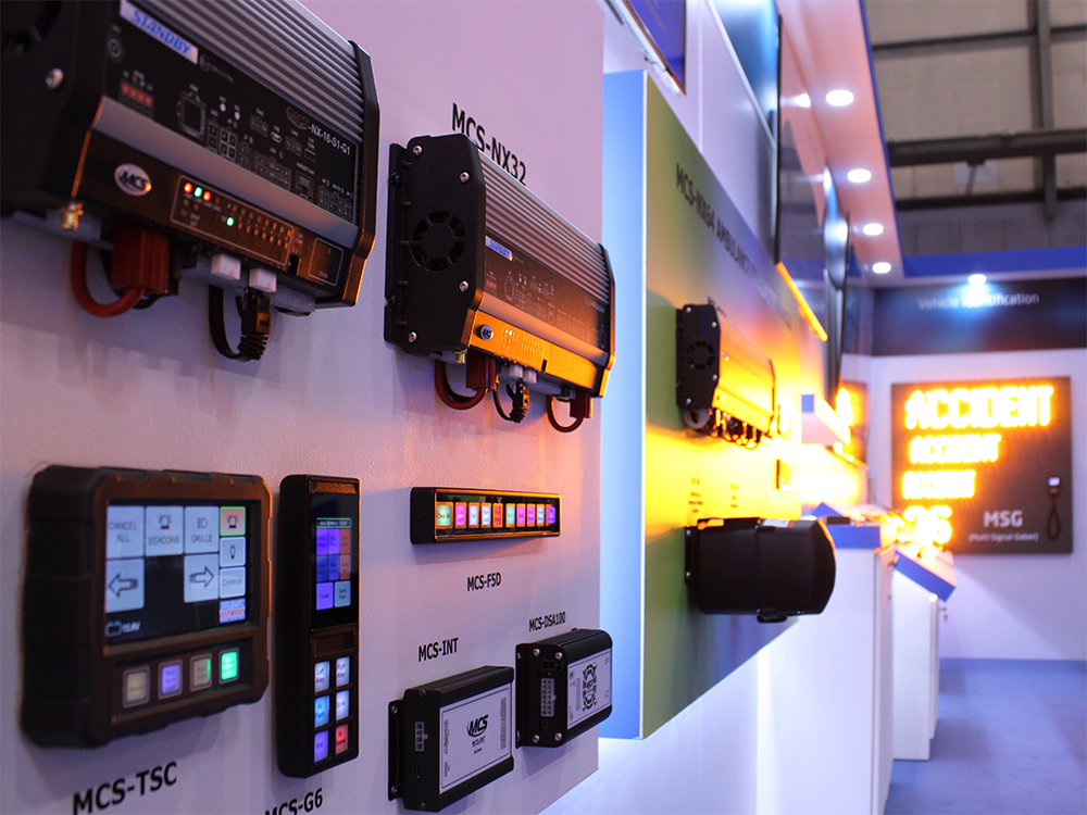 Closely cropped angle shot of various control devices mounted on the wall of an exhibition stand. A lage multi matrix display sign is lit in amber and out of focus in the background and reads @accident'.