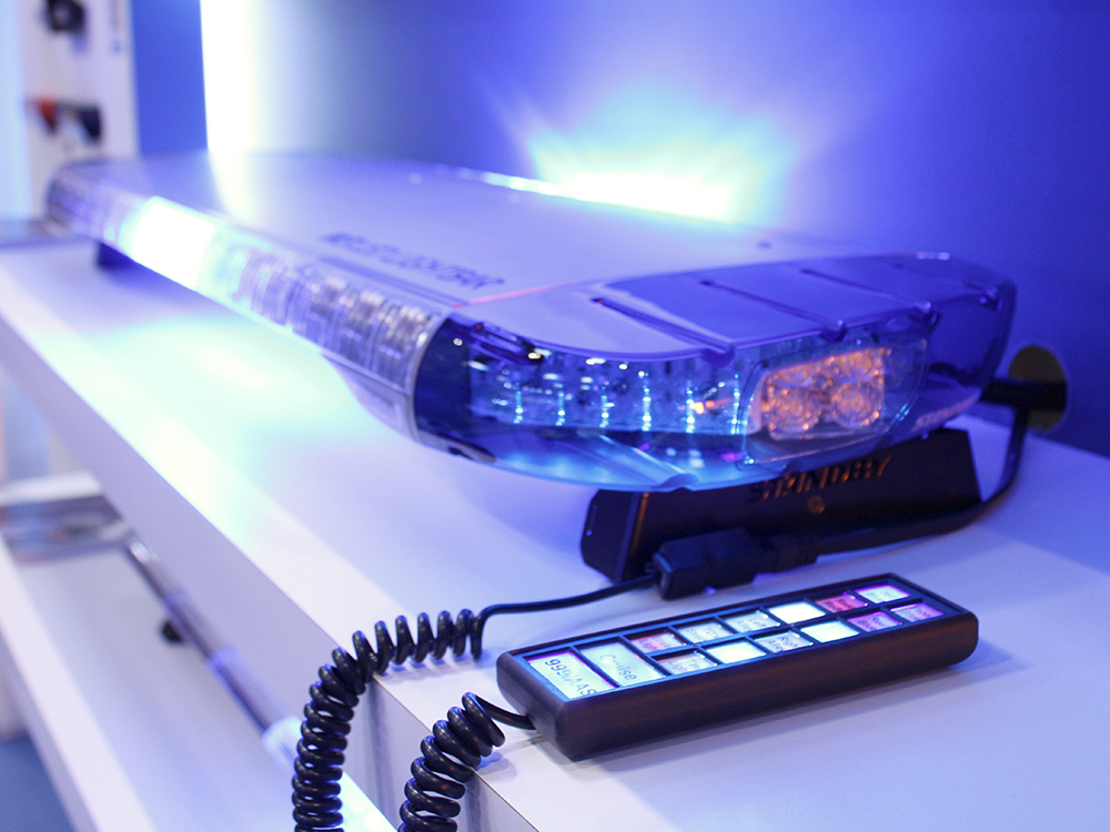 Closely cropped angle view of an emergency lightbar lit up in blue with a handset with multicolour buttons, both set on a white shelf.