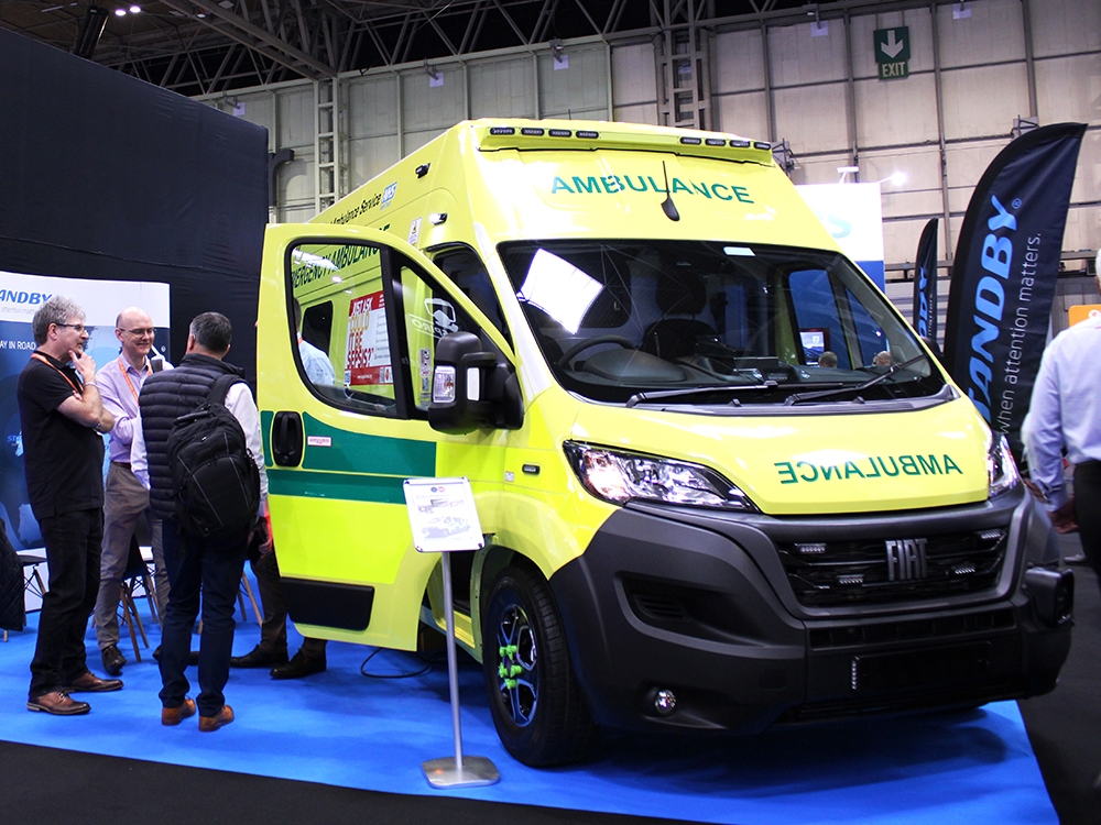 An ambulance takes up most of this image, set at an angle with it's front door open, displayed on an exhibition stand at The Emergency Services Show 2024. A group of people stand talking to the left, in front of a banner which reads 'Standby'. Two flag banners can also be seen to the right behind the vehicle which also have the Standby logo.