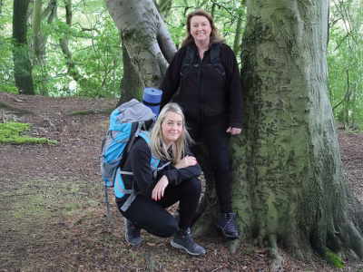 Two female Standby UK employees stand in front of a tree in the woodland. One is crouching and has a backpack with roll mat on her back.