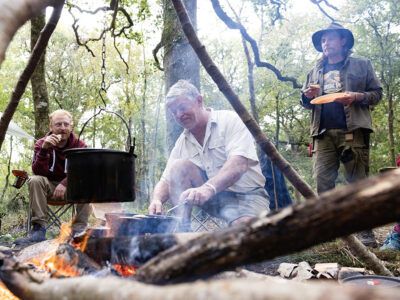 Three people gathered round a fire with a cooking pot suspended over it in the woods, during the day.