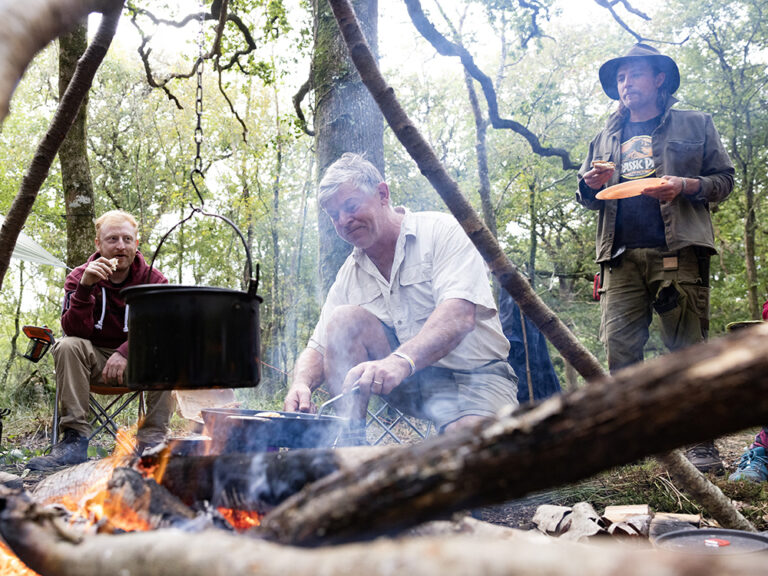 Three prople gathered round a fire with a cooking pot suspended over it in the woods, during the day.