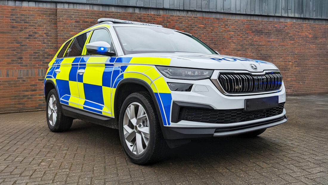 Low angle view of a police vehicle parked on paving in front of brick building. Small blue light on wing mirror is lit.