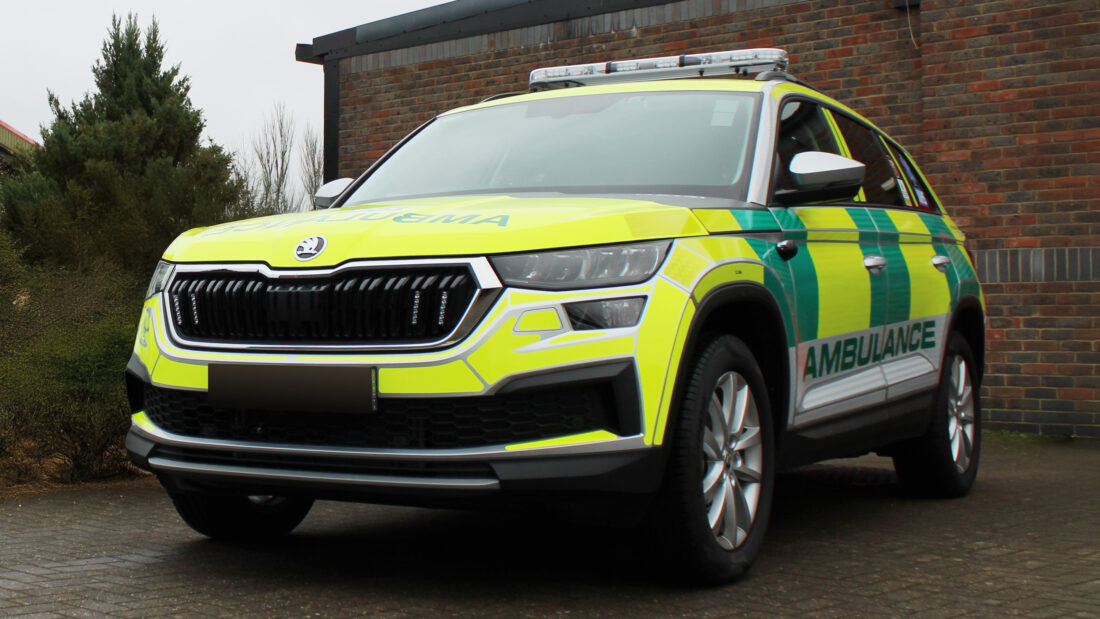 Ambulance vehicle parked at an angle in front of a brick building with foliage. Photo taken from a low angle.