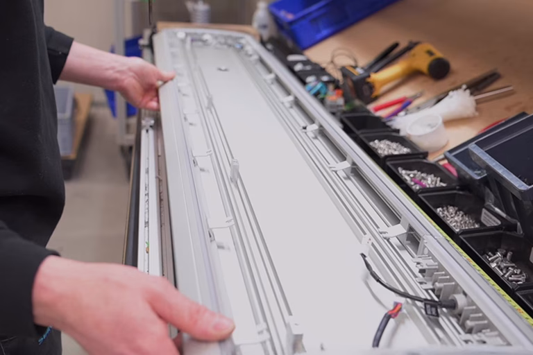Hands holding aluminium lightbar housing on workbench surrounded by tools and fasteners.