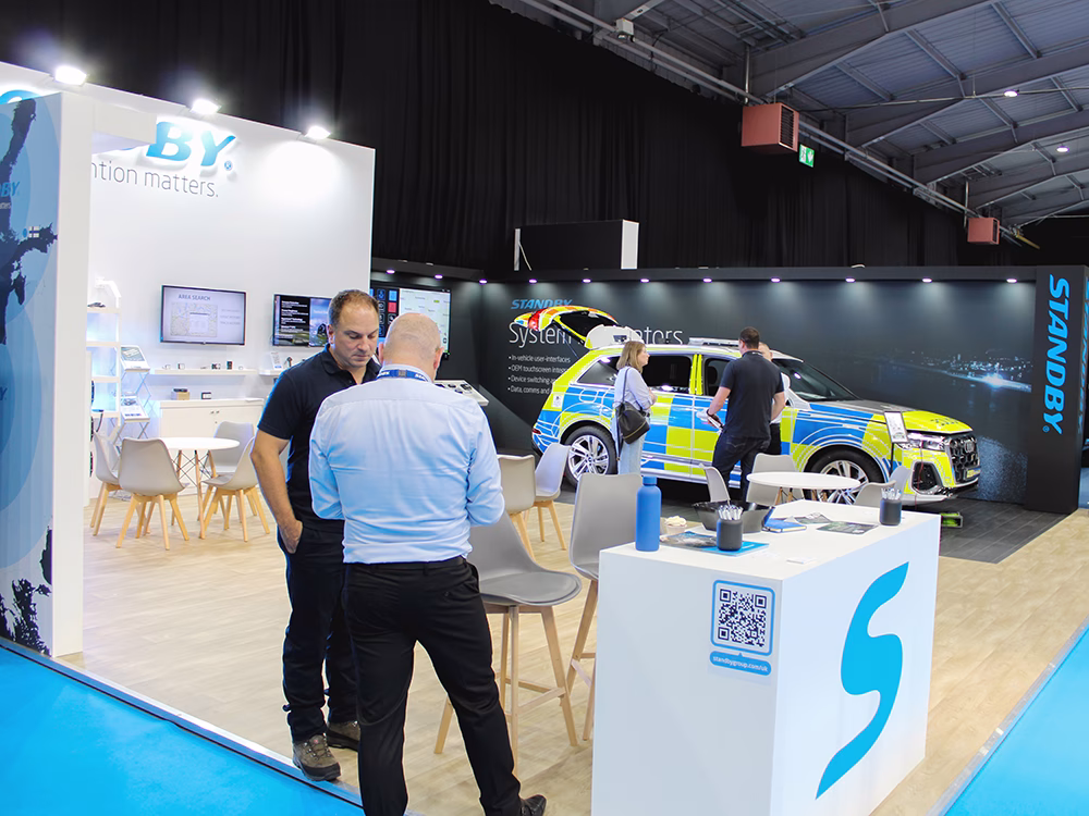 Standby UK exhibition stand at NAPFM 2025 taken at an angle, the build is modern with a black and white colour scheme and blue Standby logos, the floor is light wood effect with a darker zoned area. There are 2 clusters of stand visitors talking to Standby UK employees.