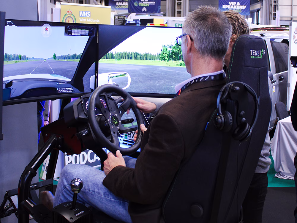 Visitor sitting in the driver's seat of the Tenstar Simulator on the Standby UK stand at the Emergency Services Show 2025, a Tenstar employee leans over to aid him, both are looking at the screens in front.