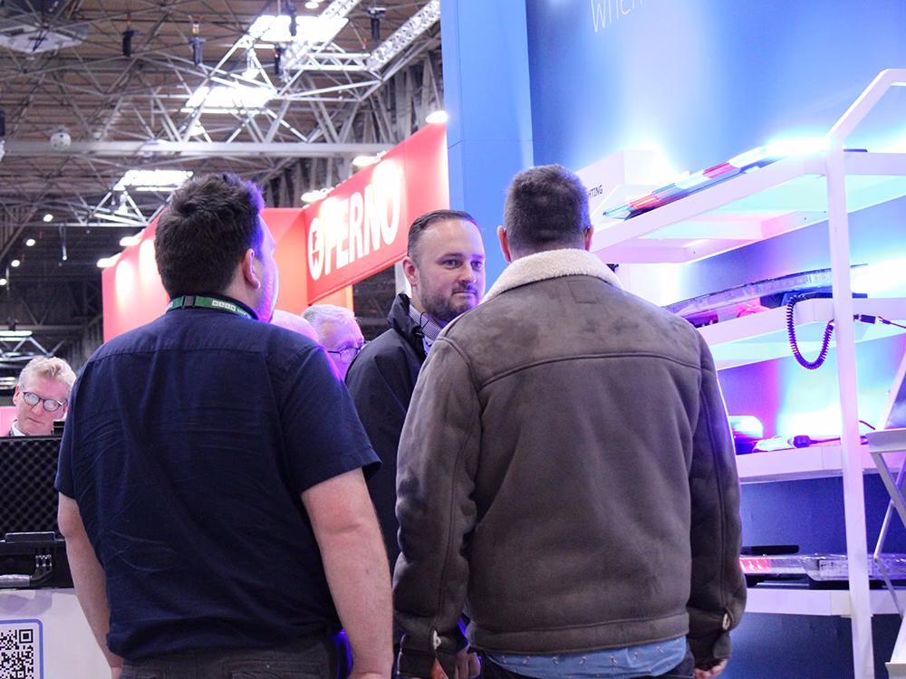 A group of three stand visitors stand in front of lightbar shelves on the Standby UK stand the Emergency Services Show 2025, two have their backs to the camera, the other seen between them in conversation.