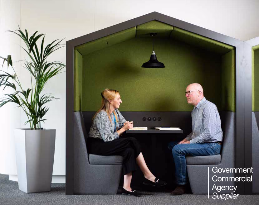 Shot of male and female CCS workers sitting across from each other in a pod with seats and table in a modern office location with potted plant. Government Commercial Agency supplier logo in lower righthand corner.