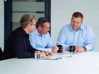Group of smartly dressed Standby employees (2 in blue shirts and one in a black jumper) sit at a white table in a sleek boardroom interacting with each other.