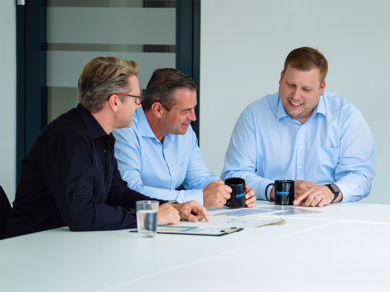 Group of smartly dressed Standby employees (2 in blue shirts and one in a black jumper) sit at a white table in a sleek boardroom interacting with each other.