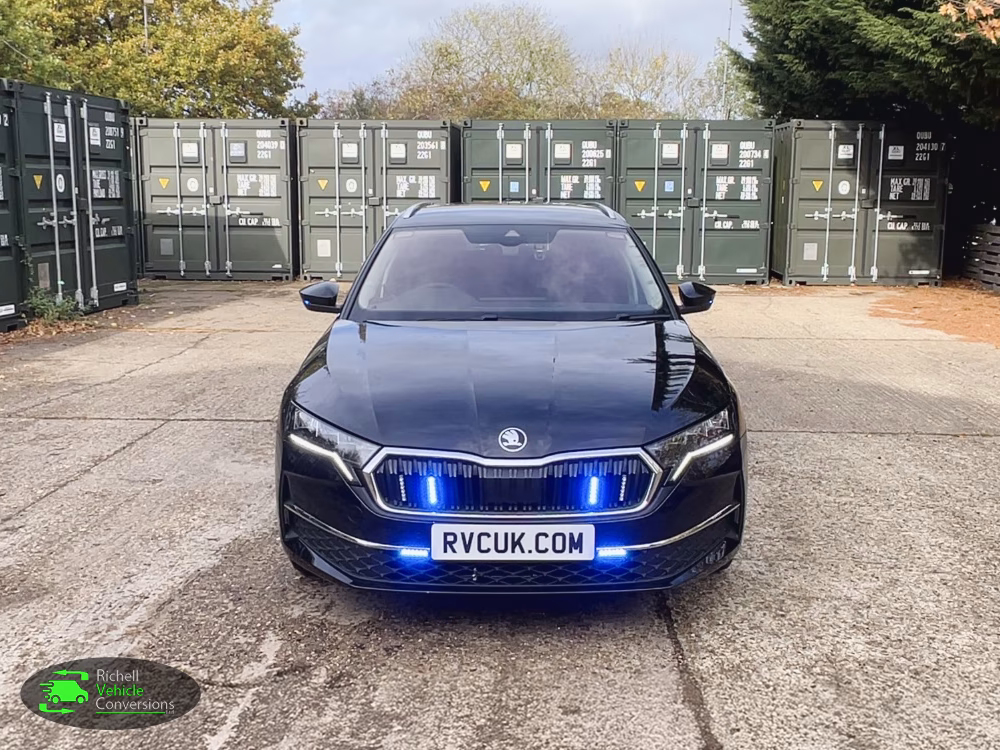 RVC Skoda Octavia Driver Training Vehicle shown from the front with blue lights parked on a concrete surface in front of large green shipping containers.