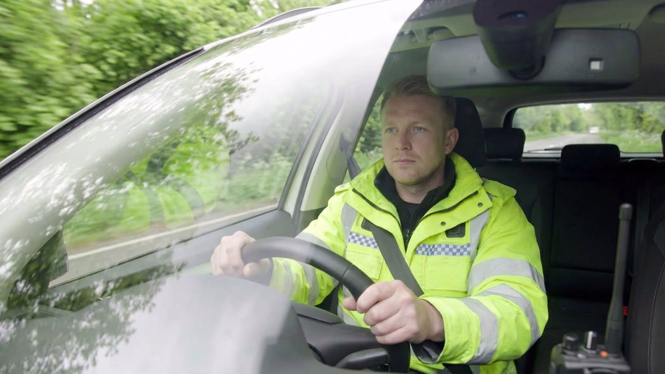 Front bonnet view of a police driver wearing a hi-vis jacket driving down a rural road, his hands are on the steering wheel and eyes on the road.