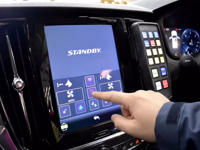 Hand with navy blue shell sleeve reaches to press a button on the Volvo infotainment screen CARAT interface. A tactile Standby handset is also mounted alongside this.