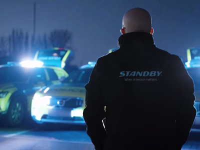 Standby employee stands with his back to the camera, wearing a Standby branded jacket with the collar up, in front of three Police vehicles out of focus in the background. The photo is taken at night.