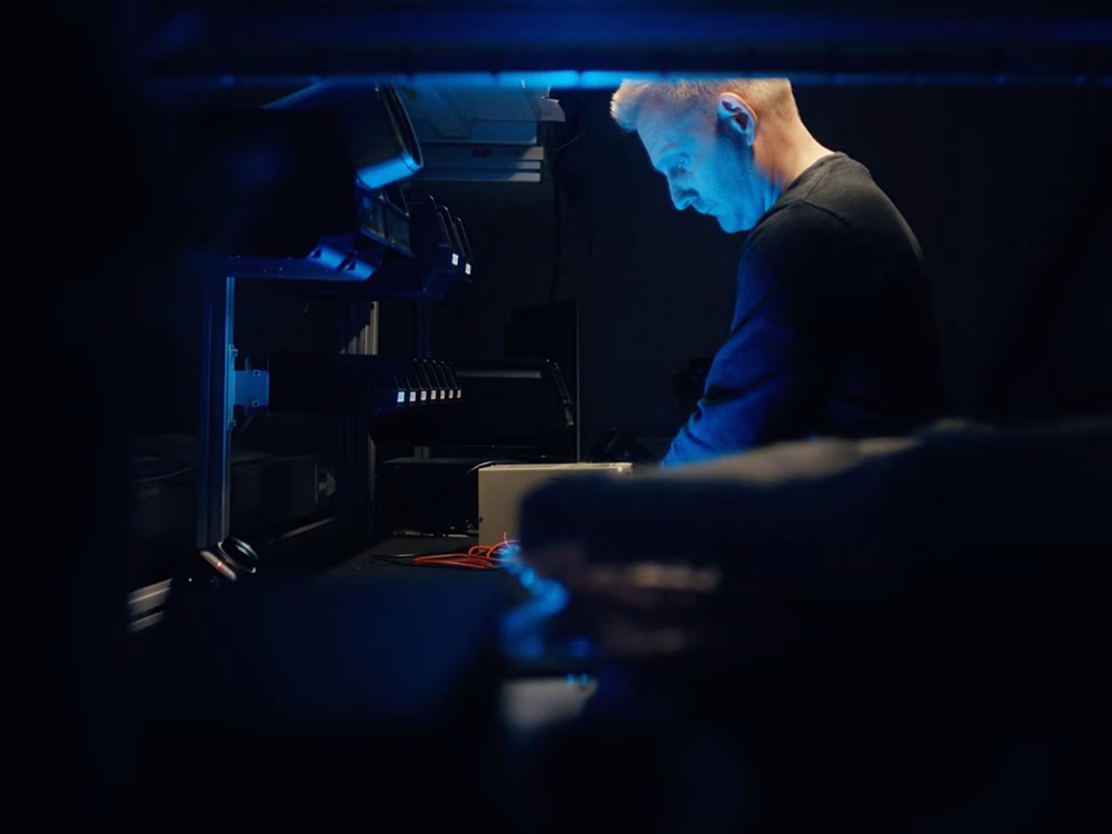 Engineer in a dark room working at a bench, standing side on and illuminated by a blue light, equipment silhouetted in the foreground, out of focus
