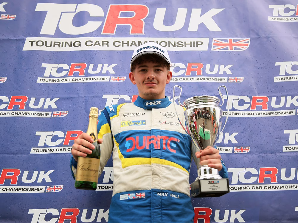 Max Hall stands with cup and champagne in front of TCR UK branding in racing suit
